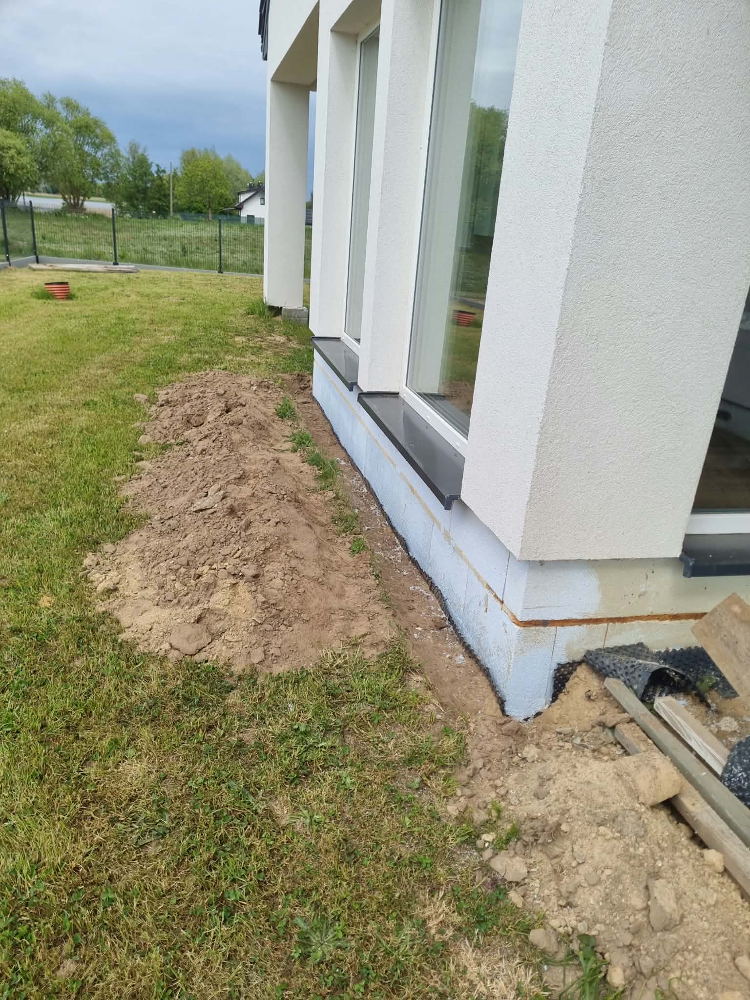 Jasna struktura elewacji i ciemne parapety; strefa cokołu przed końcowym wykończeniem.